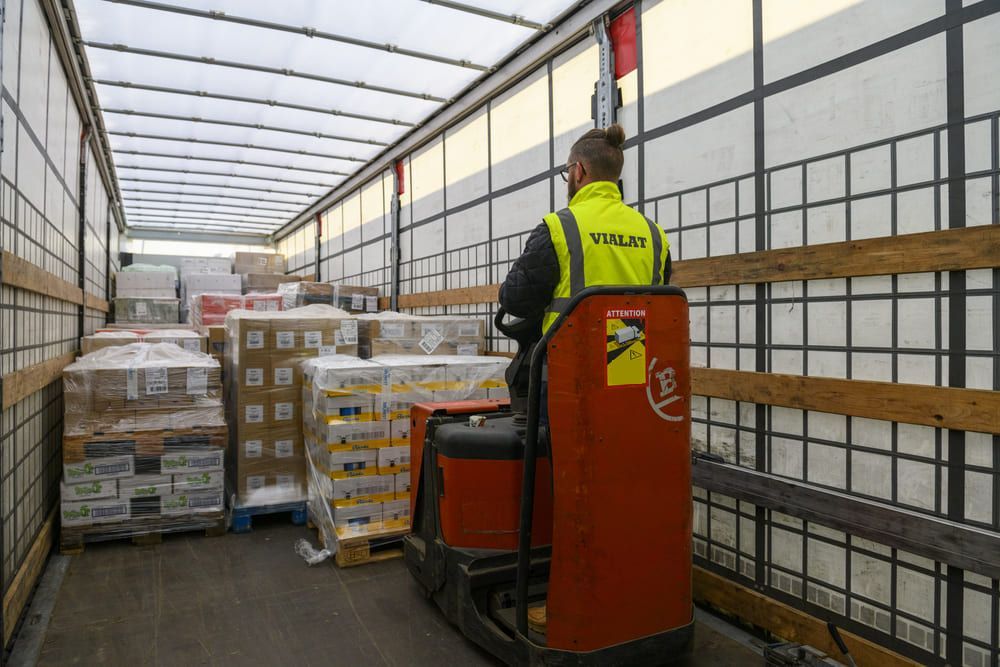 Un ouvrier conduit un chariot élévateur rouge dans une allée d'entrepôt bordée de palettes empilées.