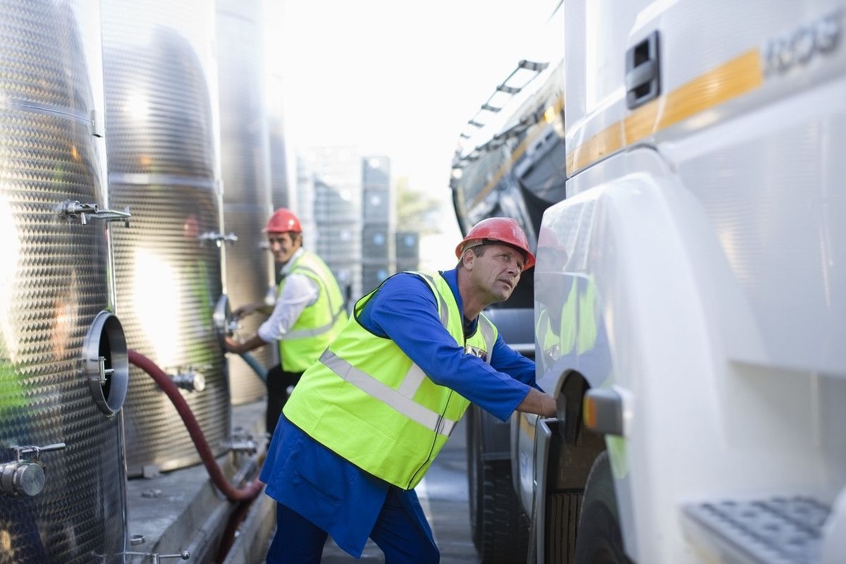 Des ouvriers portant des gilets de sécurité fluo et des casques rouges nettoient un camion-citerne blanc dans une zone industrielle.