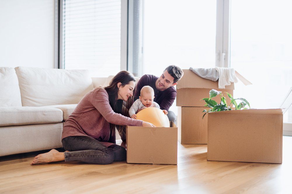 Une famille déballe des cartons dans un salon lumineux, avec un bébé entre deux adultes.