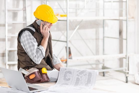 Un trabajador de la construcción está sentado en el suelo hablando por un teléfono celular mientras mira una computadora portátil.