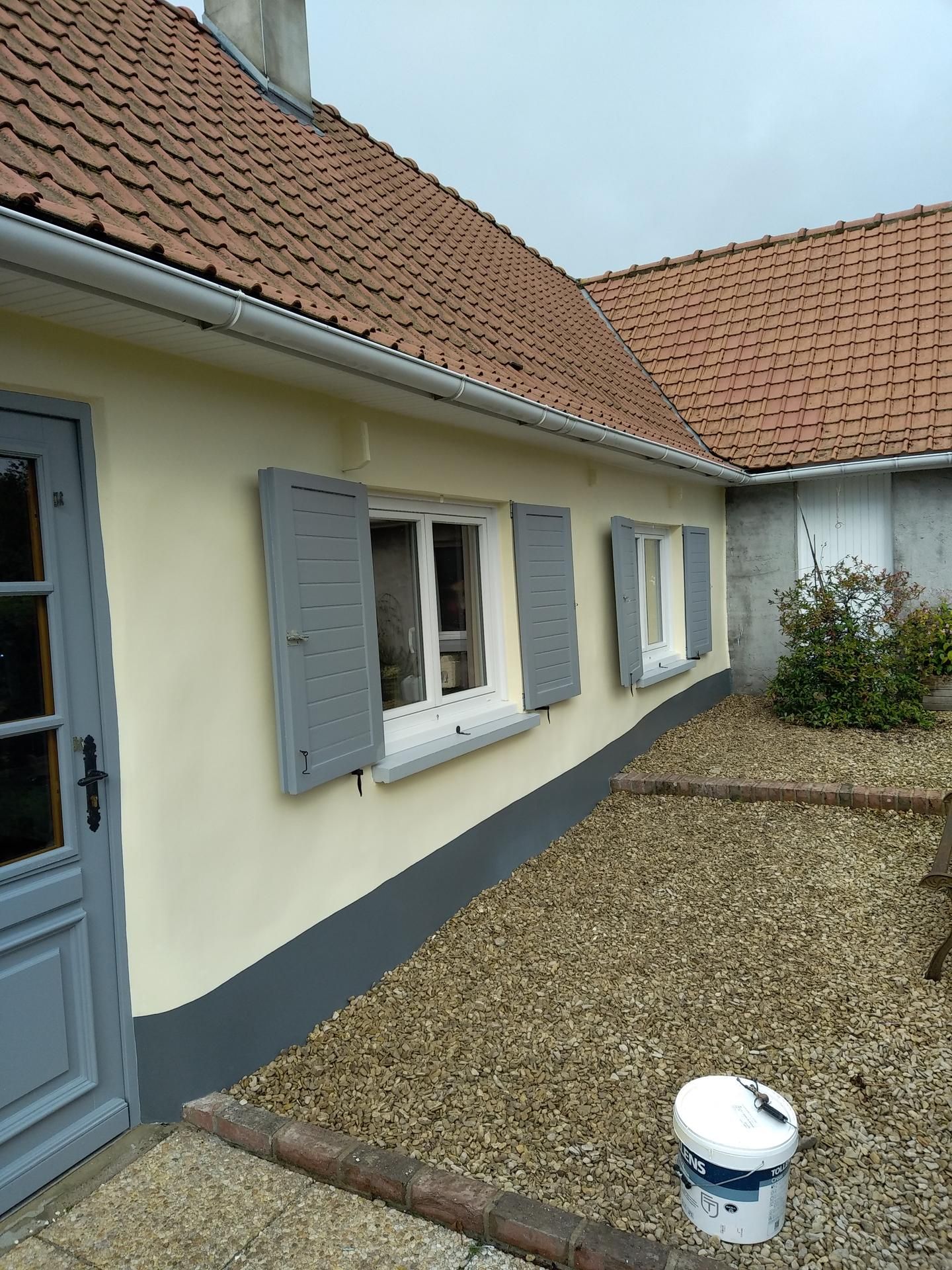 Un cottage couleur crème avec des volets gris et une porte grise assortie, doté d'un toit de tuiles et d'une cour avant en gravier.