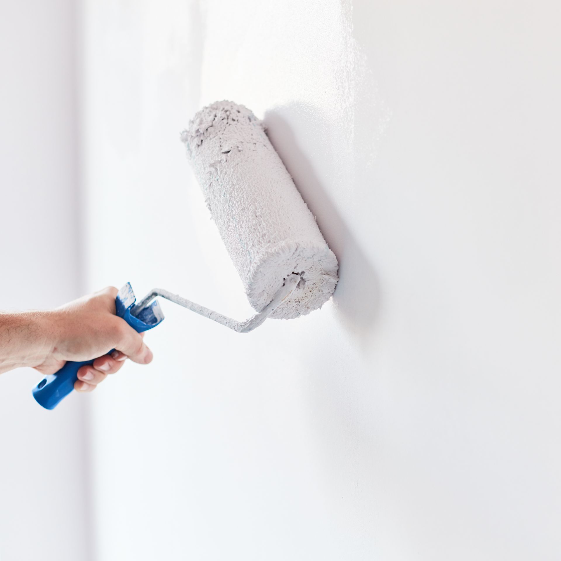 Une main tenant un rouleau à peinture bleu, appliquant une couche de peinture blanche sur un mur blanc vierge.