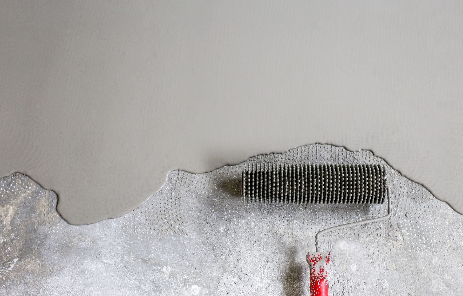 Un rouleau à pointes se déplace sur une surface grise humide, éliminant les bulles d'air d'un revêtement béton fraîchement appliqué.