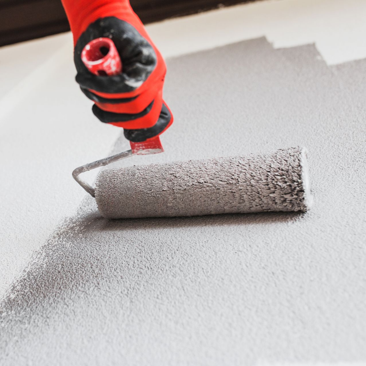 Une personne portant des gants rouges utilise un rouleau à peinture pour appliquer de la peinture grise sur une surface.