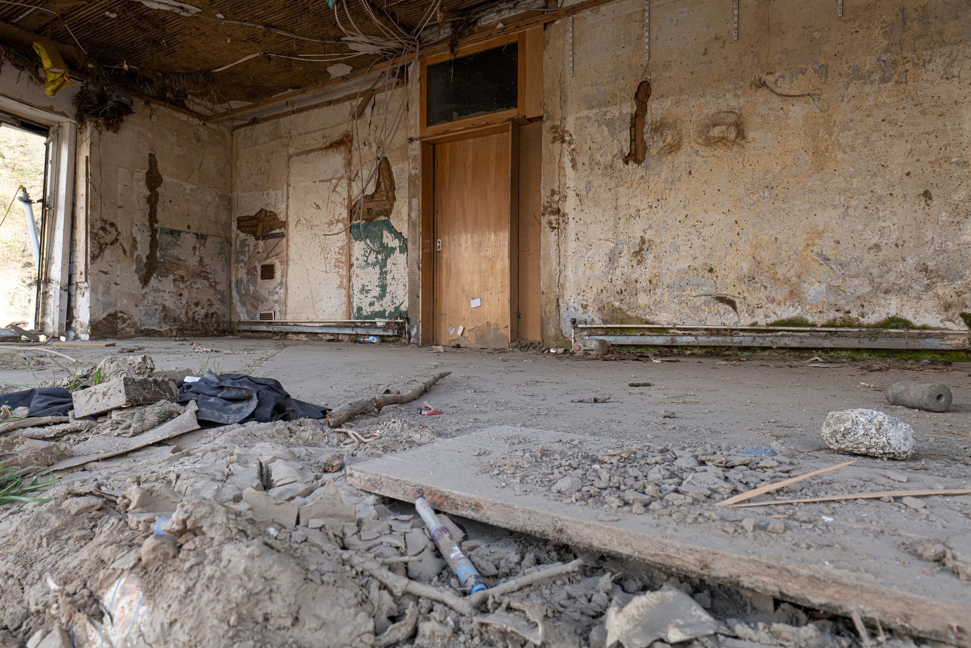 Une pièce délabrée aux murs qui s'effritent, au sol jonché de débris et dotée de deux portes en bois fermées.