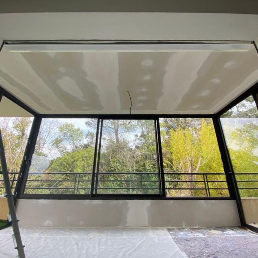 Une pièce en rénovation avec des plafonds et des murs en plaques de plâtre, dotée de grandes portes coulissantes en verre donnant sur une forêt.