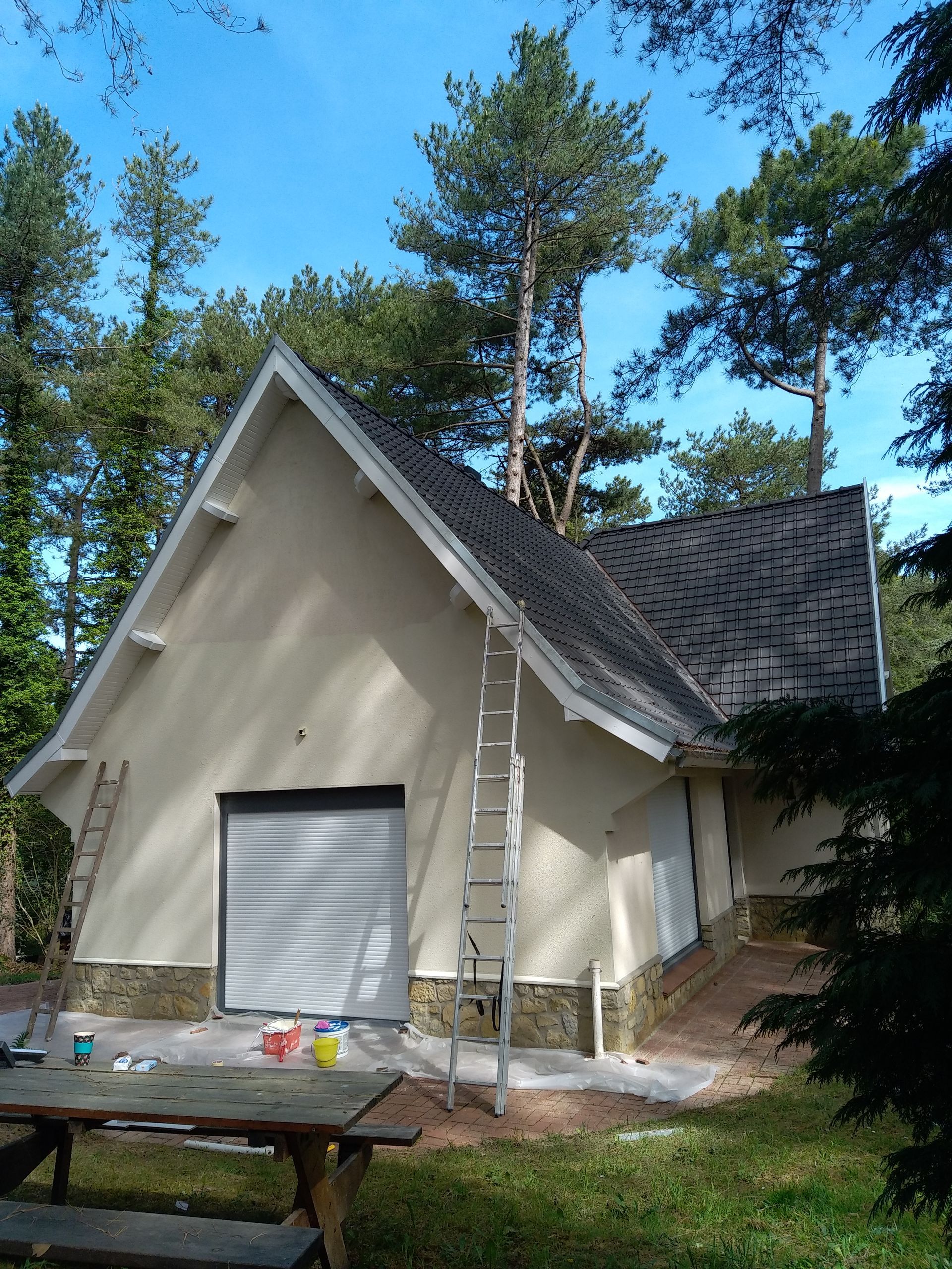 Un bâtiment clair en forme de A, sous des arbres, avec des échelles appuyées contre ses murs, lors de travaux de peinture extérieure.