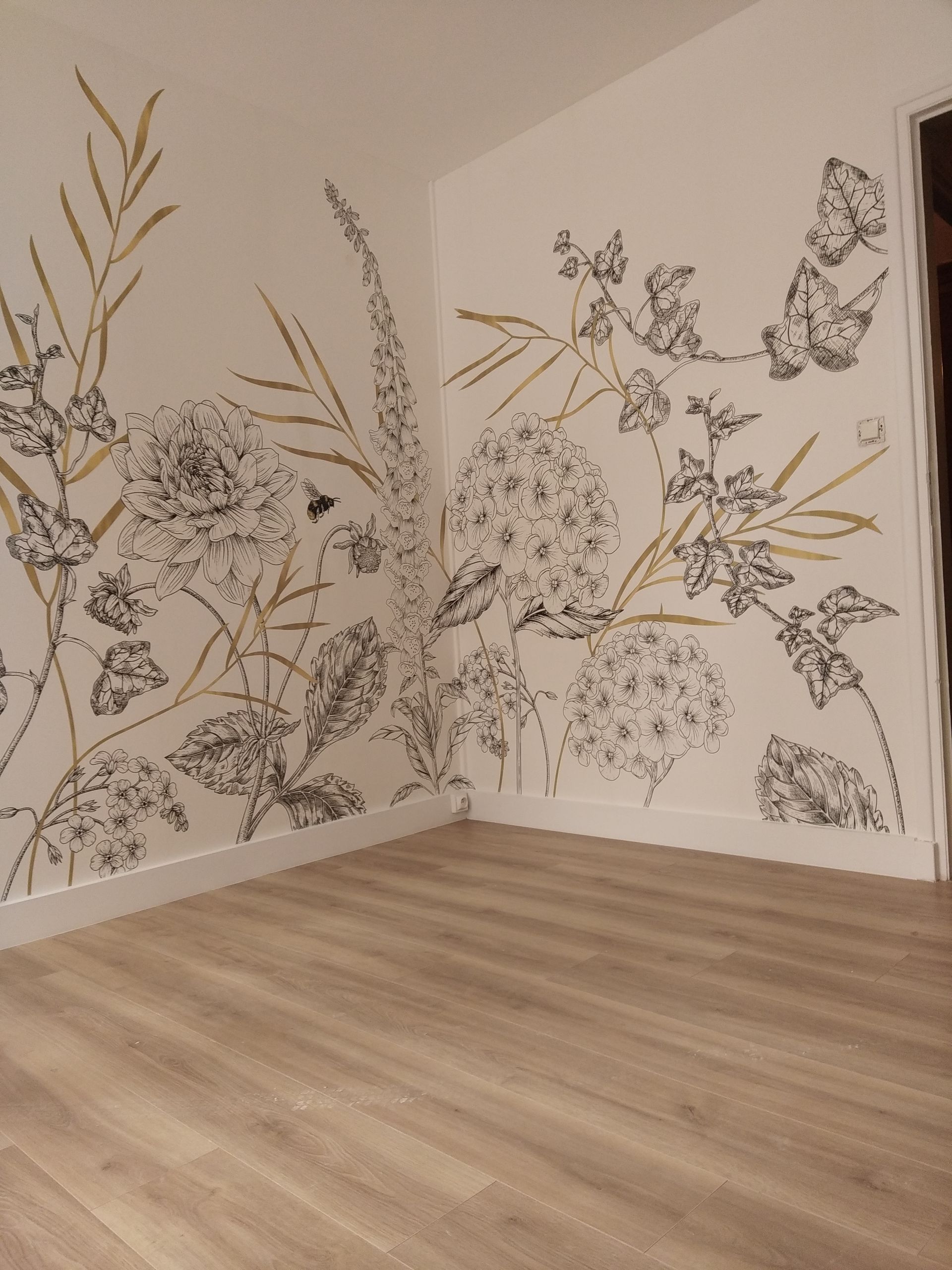 Un coin de pièce orné d'une délicate fresque murale florale dessinée à la main, à l'encre noire et à la peinture dorée sur des murs blancs.