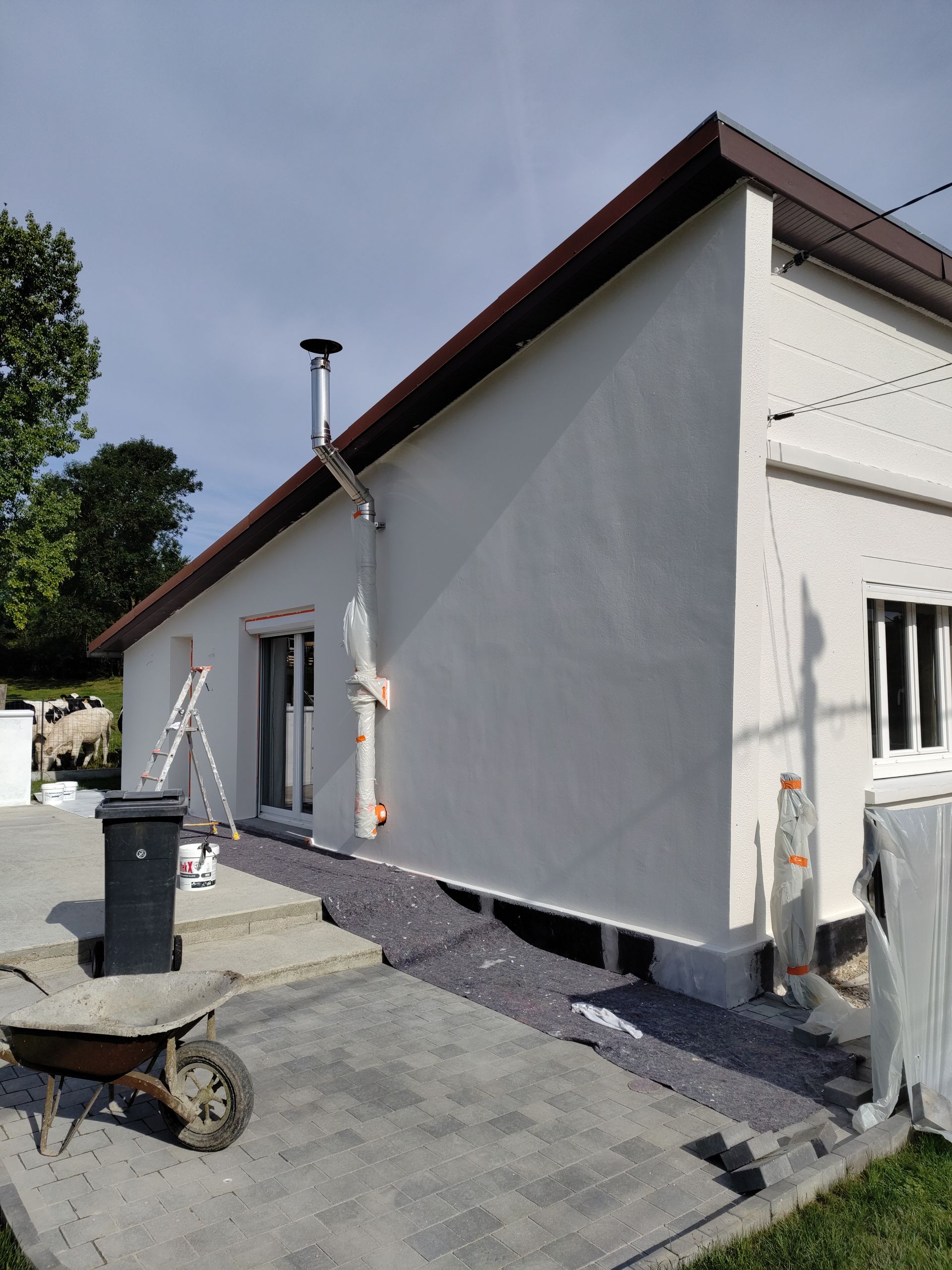 Une maison blanche au toit en pente et à la conduite d'eau neuve, située à côté d'une terrasse avec du matériel de chantier et du gravier.