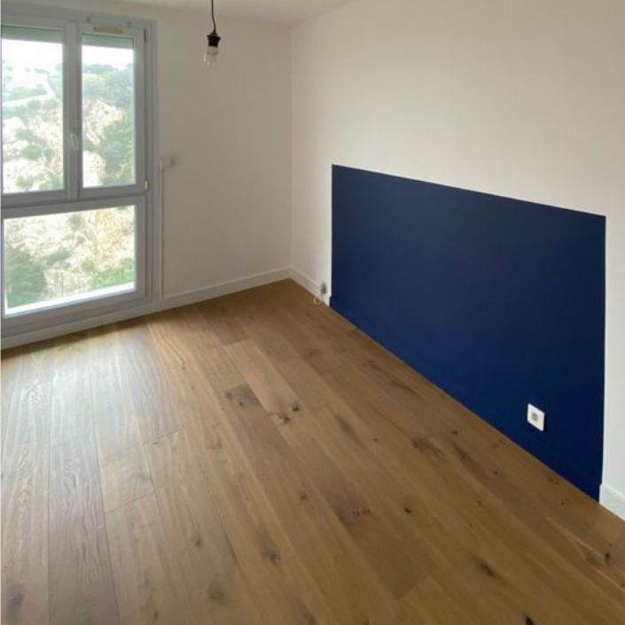 Une pièce vide avec un parquet clair, des murs blancs et une grande fenêtre, agrémentée d'un pan de mur peint en bleu foncé.