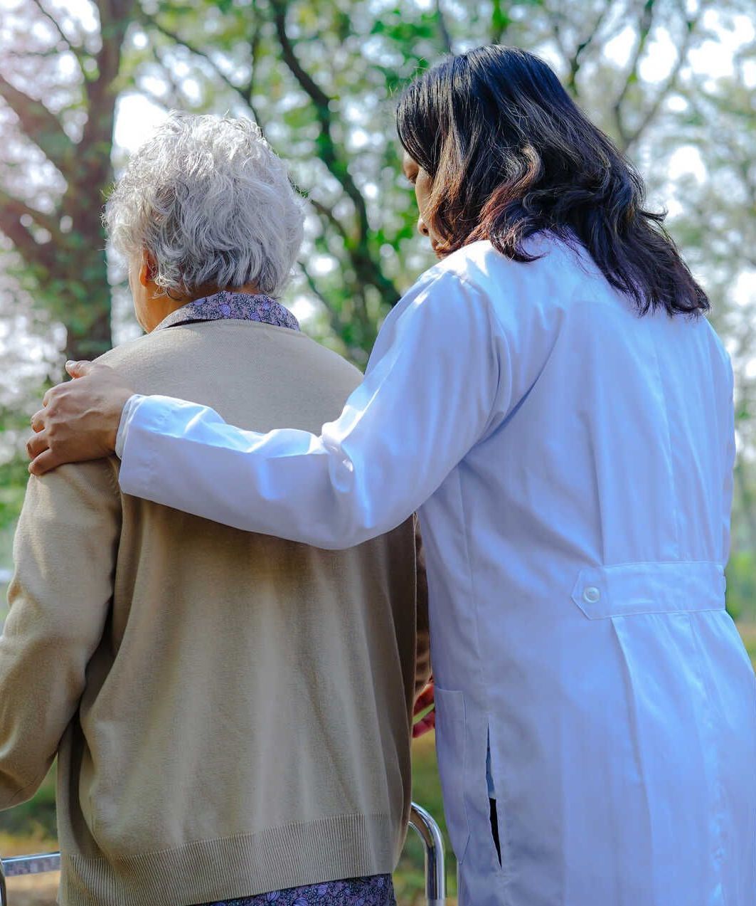 Eine Frau im weißen Kittel hilft einer älteren Frau im Rollstuhl.