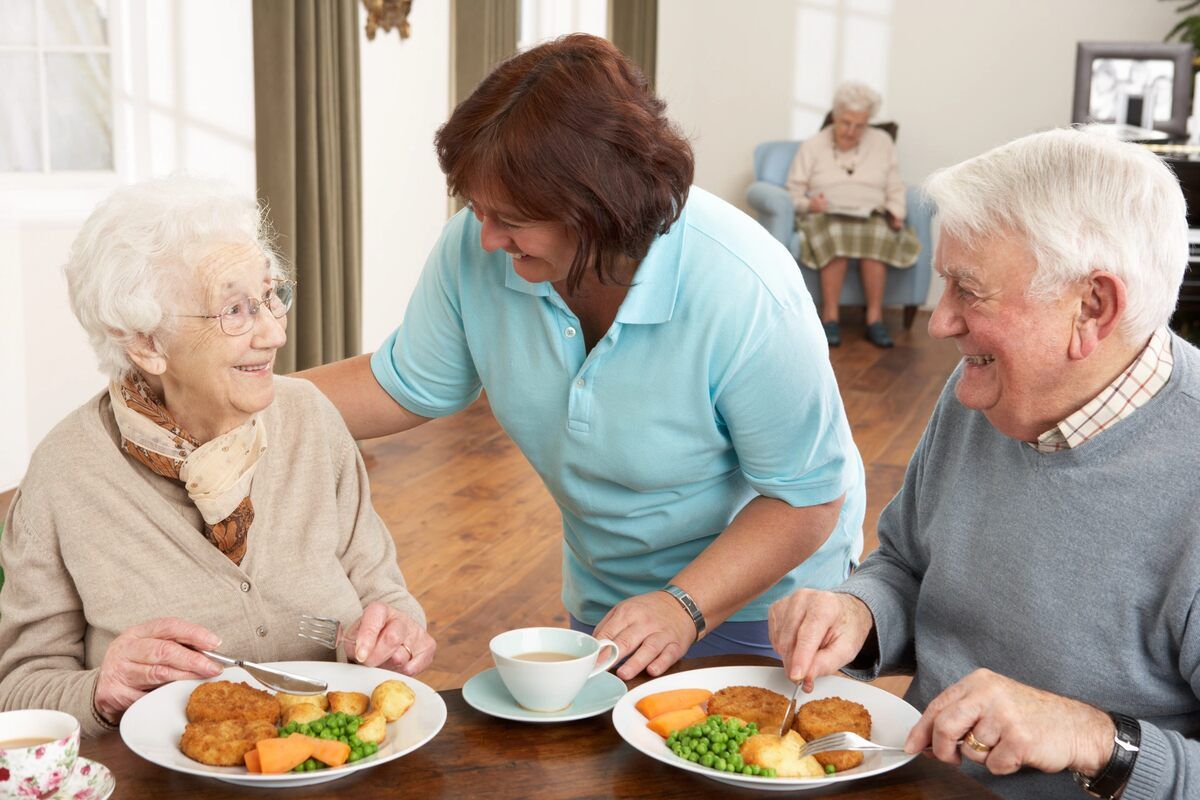 Eine Frau serviert zwei älteren Menschen an einem Tisch Essen.