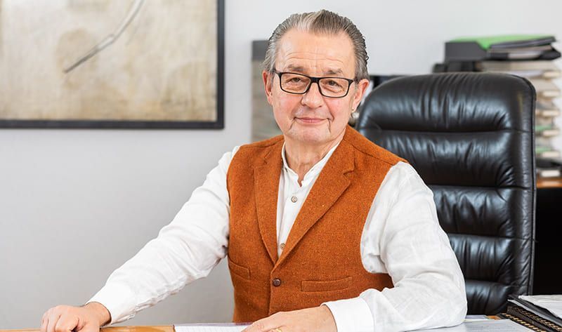 Ein älterer Mann mit Brille und Weste sitzt an einem Schreibtisch.