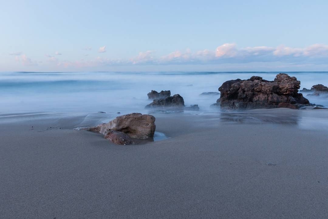 Una fotografía de larga exposición de una playa de arena con rocas en primer plano y el océano en el fondo.