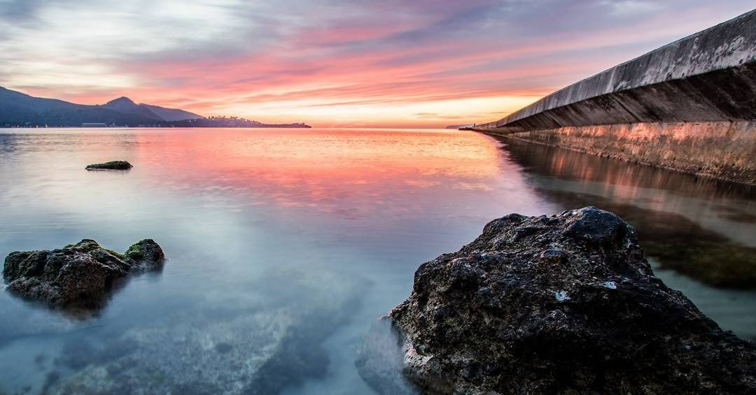 Una puesta de sol sobre un lago con rocas en primer plano y montañas al fondo.