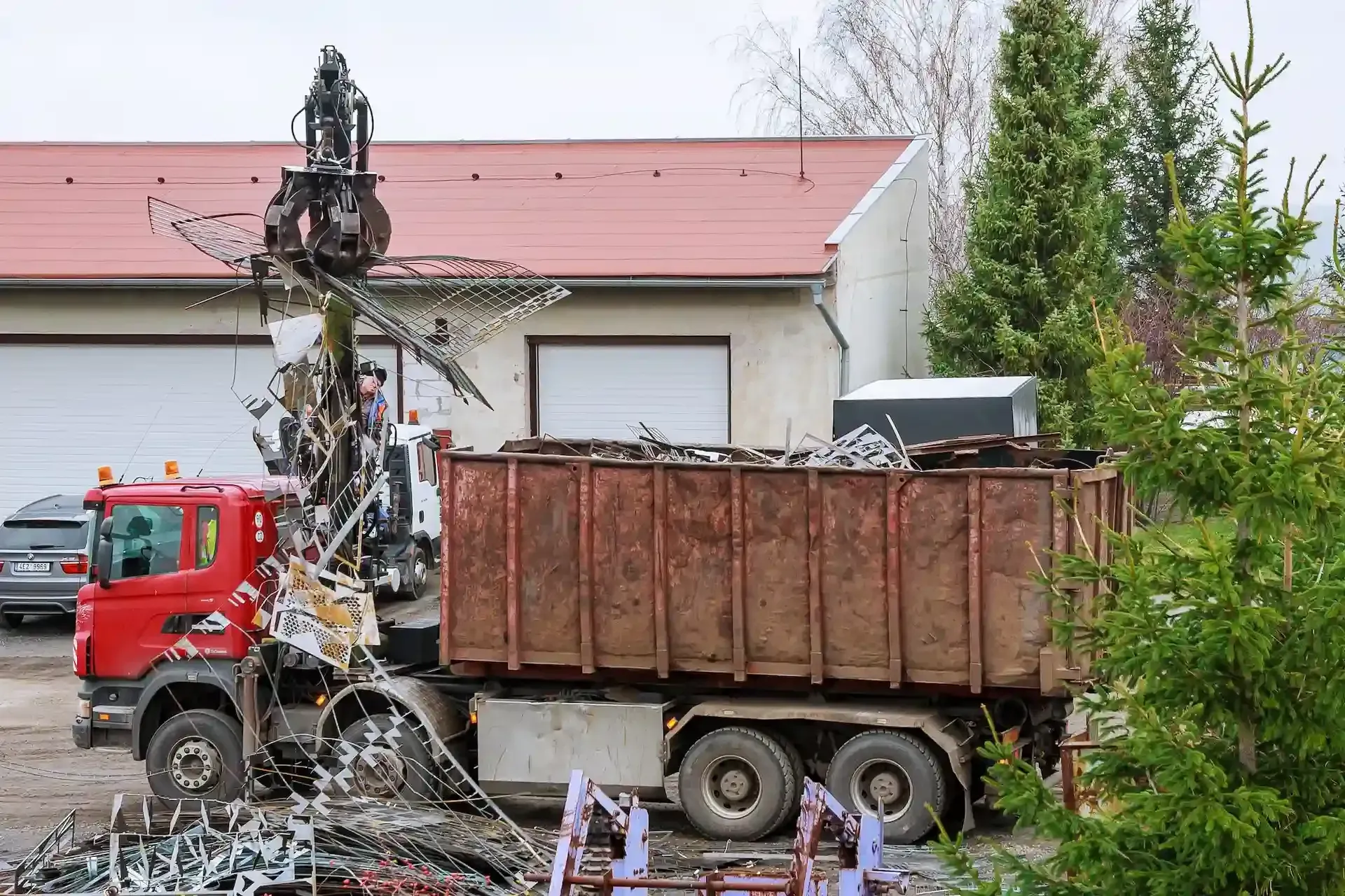 Camión rojo cargando chatarra en un contenedor grande. Se utiliza un mecanismo similar a una garra.