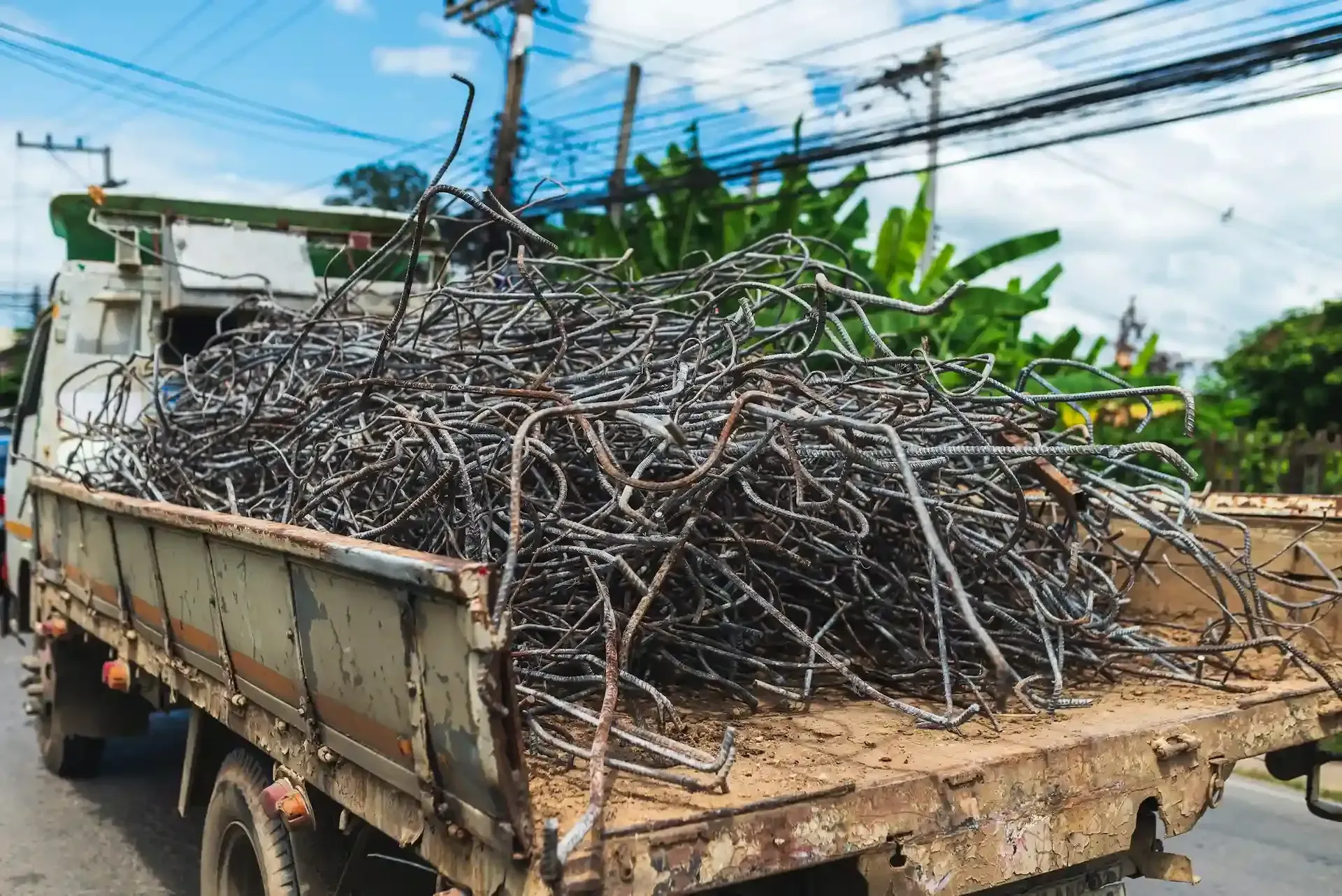 Caja de camión llena de chatarra en una calle con líneas eléctricas y follaje verde al fondo.