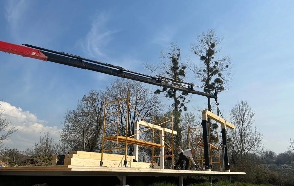 Grúa levantando vigas de madera durante la construcción de una casa al aire libre en un día soleado.