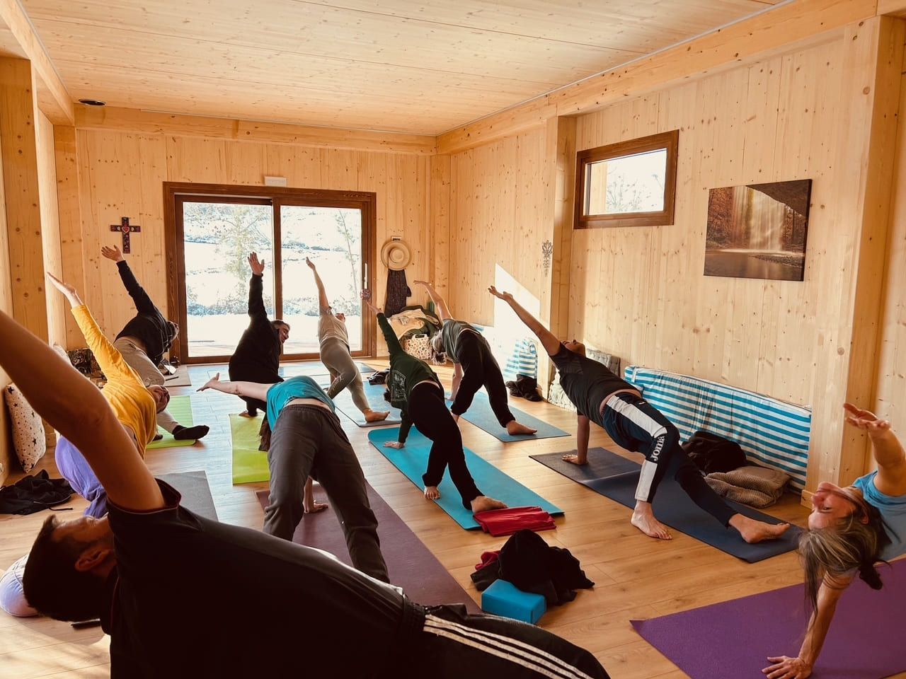 La gente practica yoga en una sala con paneles de madera. Diversas posturas, luz natural y colchonetas visibles.