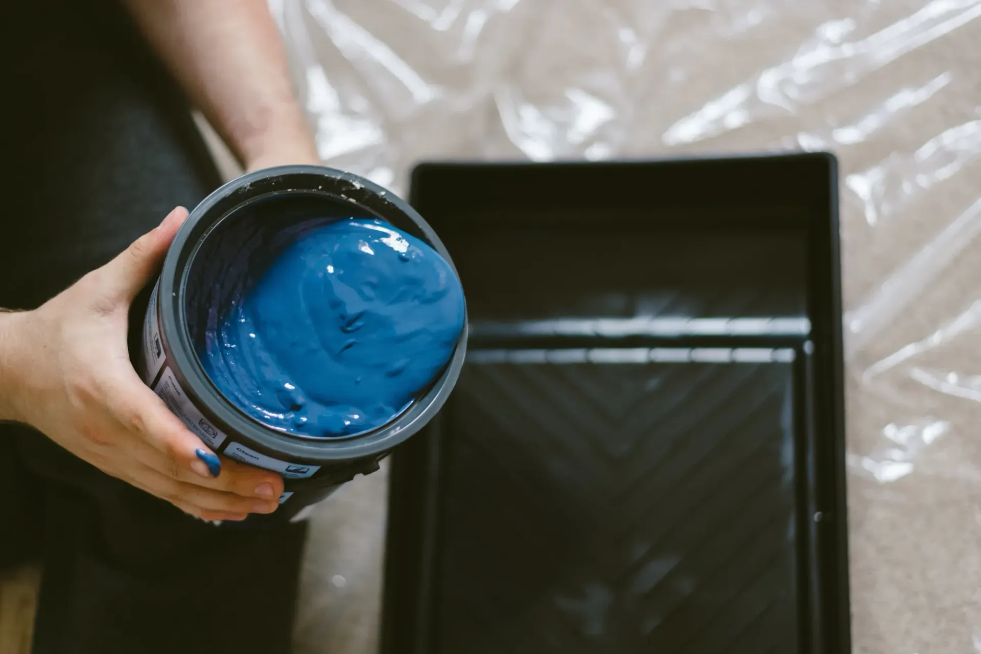 Una persona sostiene una lata de pintura llena de pintura azul vibrante sobre una bandeja de pintura negra vacía.
