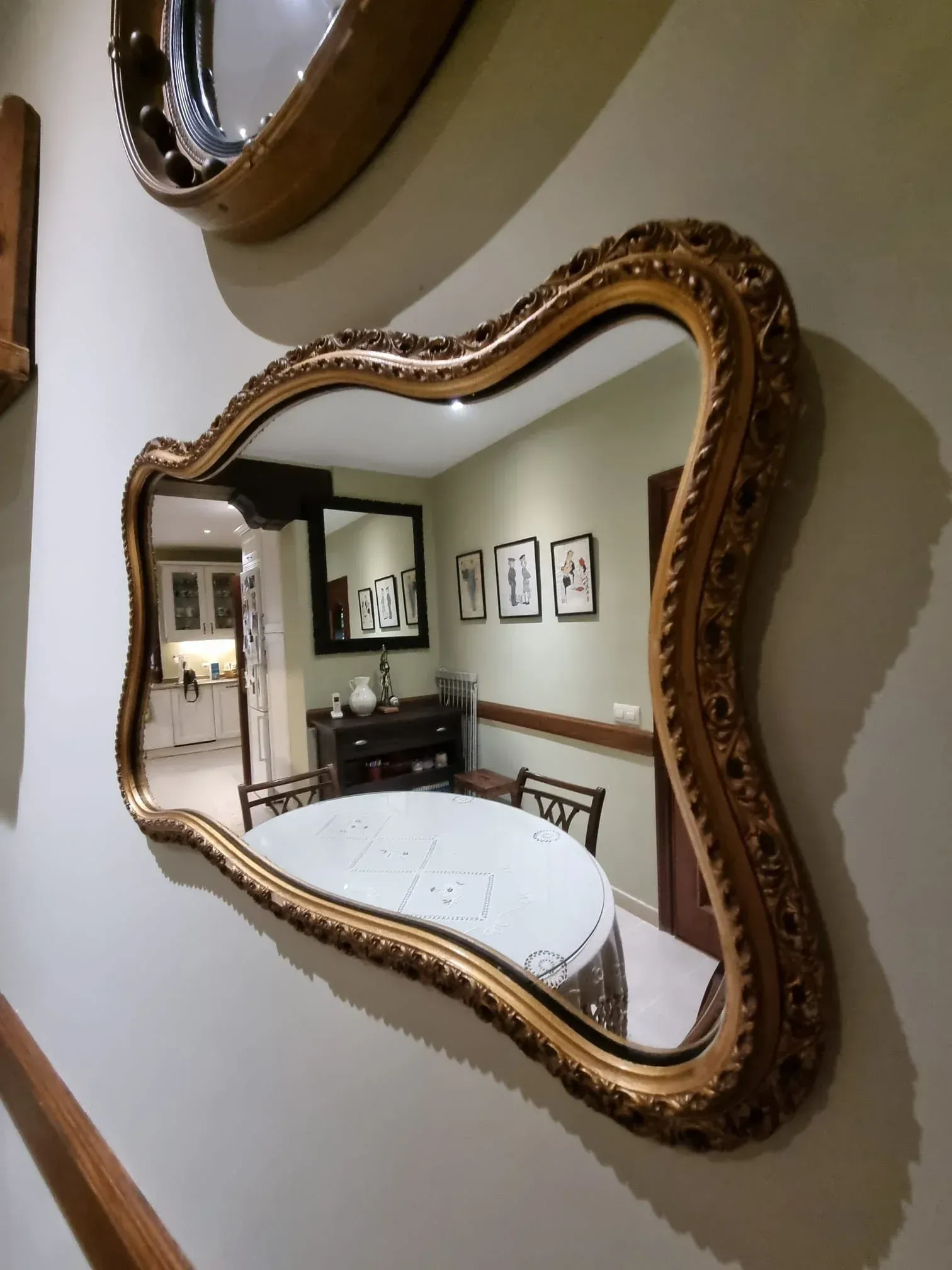 Un espejo de pared decorativo con marco dorado ondulado que refleja un comedor con una mesa blanca y arte enmarcado.
