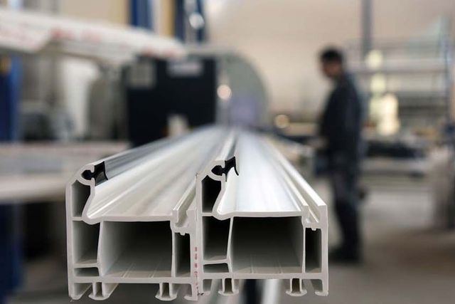 Primer plano de perfiles de ventanas de PVC blanco en una fábrica, con un trabajador borroso al fondo.