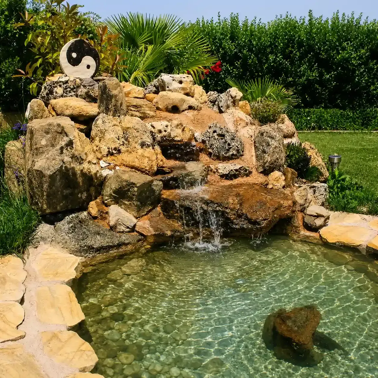Cascada de roca que cae en un estanque cristalino en un jardín exuberante con un adorno de yin-yang en las rocas.