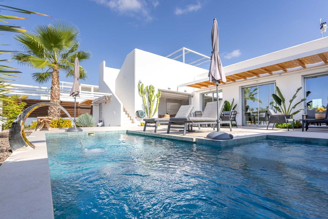 Poolside view of a white villa with a pool, palm trees, and lounge chairs under a blue sky.
