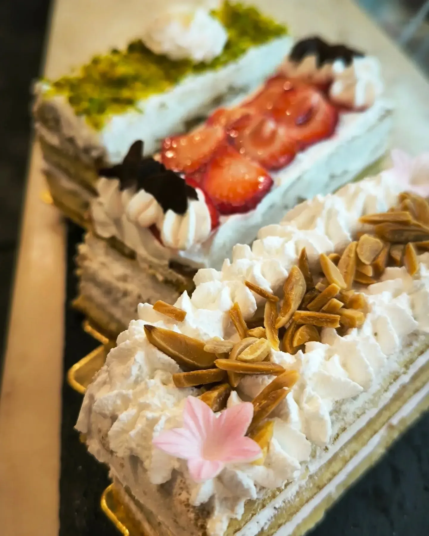 Tres rebanadas de pastel con crema batida, fresas y almendras sobre una mesa.