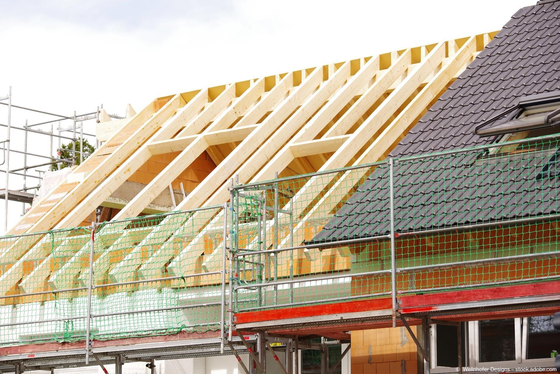 Baustelle mit einem Dach aus Holzrahmen. Darunter sind ein Gerüst und ein teilweise fertiges Gebäude zu sehen.