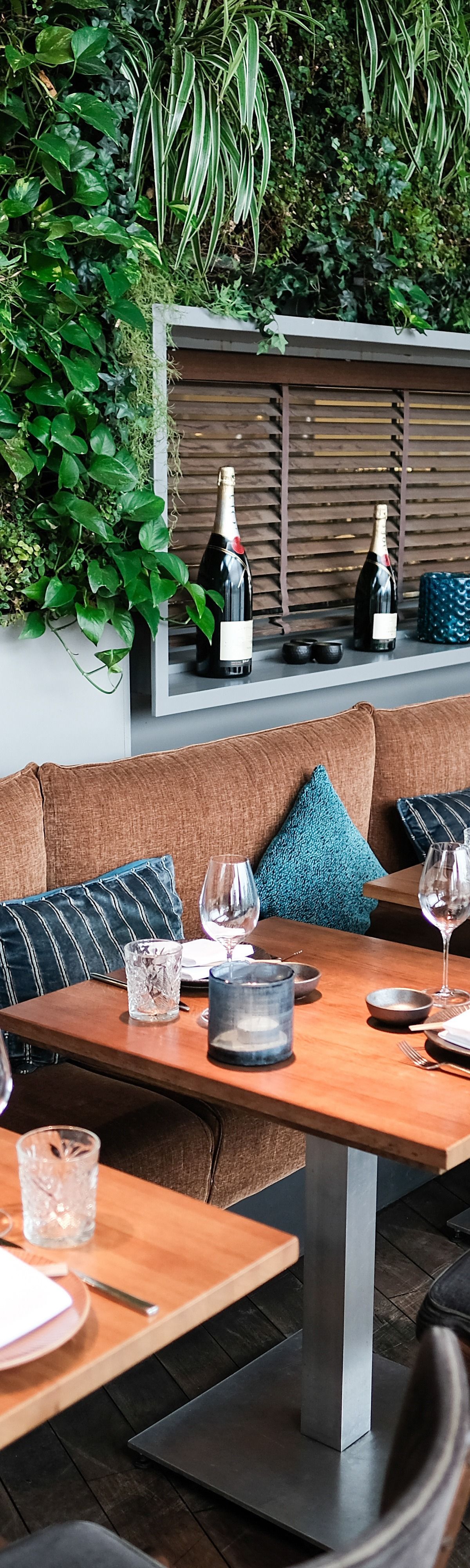 Restaurant avec tables en bois, coussins et verdure.