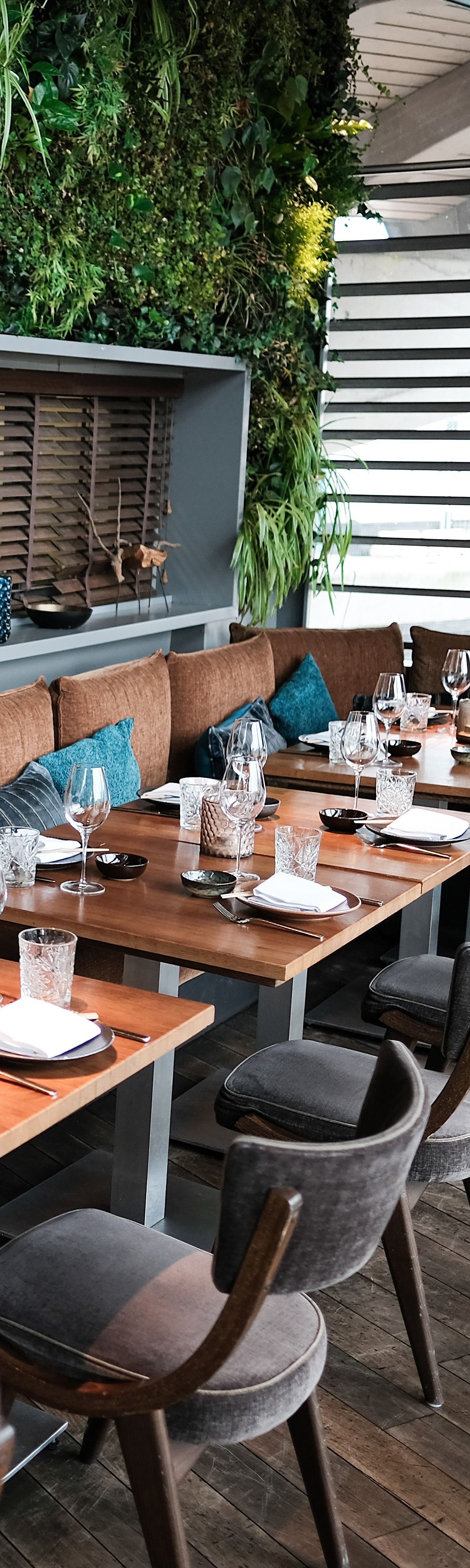 Sièges de restaurant avec tables en bois, chaises rembourrées grises et un mur de verdure.