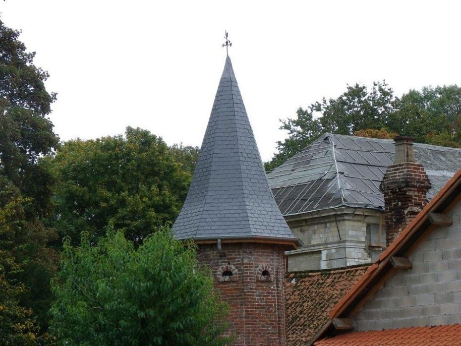 Tour en briques avec un toit pointu en ardoise, une girouette et des arbres environnants.