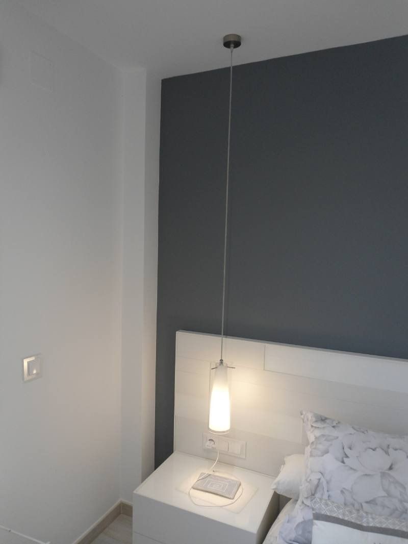 Un dormitorio con cama, mesita de noche, lámpara y pared gris.