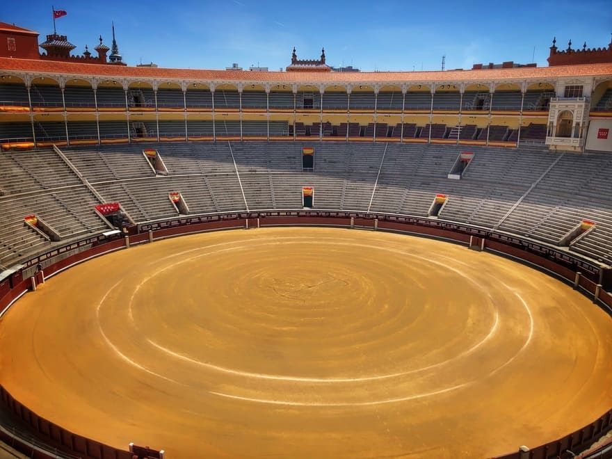 Una plaza de toros vacía con una valla roja y un cielo azul al fondo.