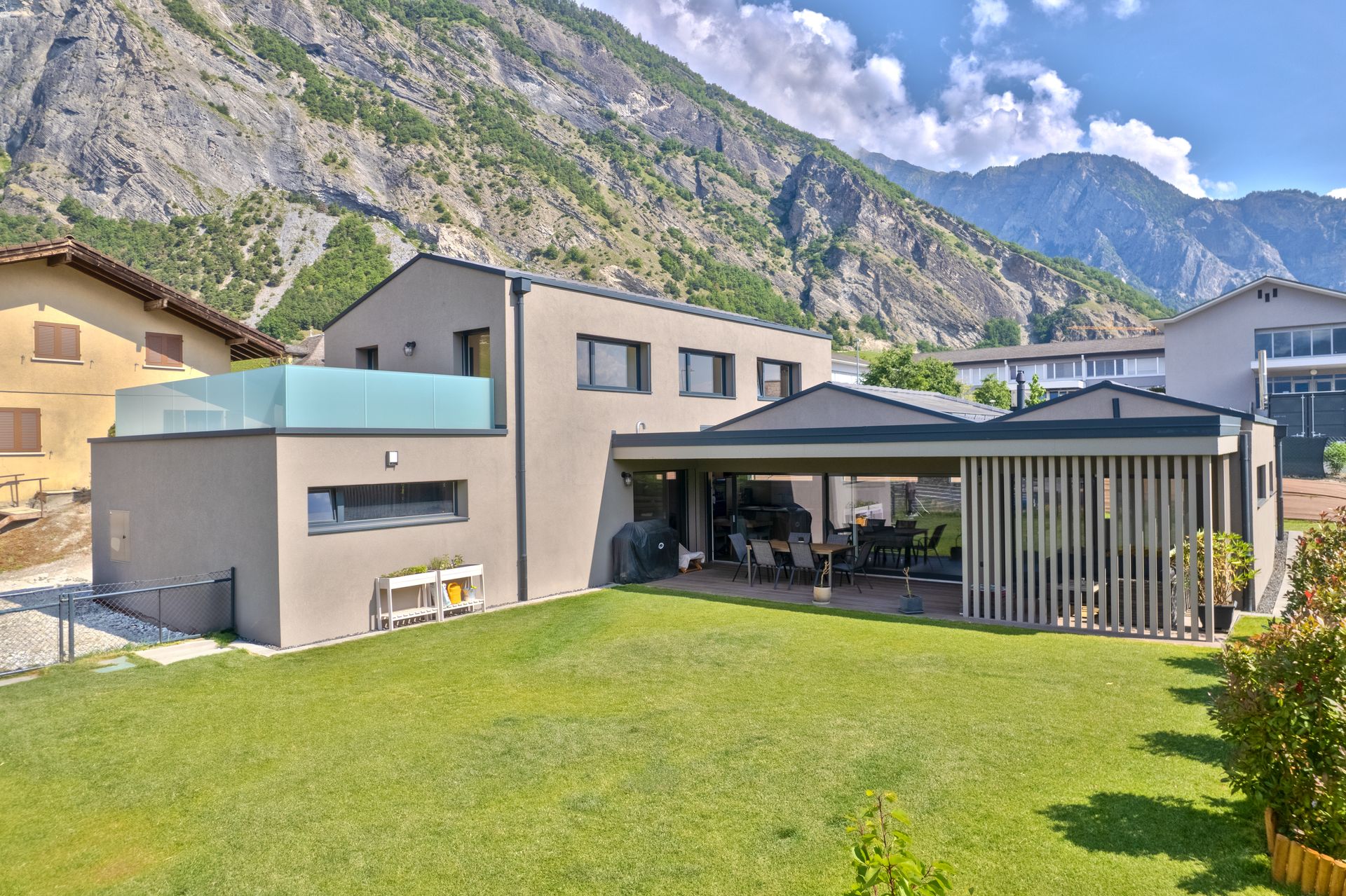 Maison moderne avec façade grise, pelouse verte, patio et vue sur les montagnes.