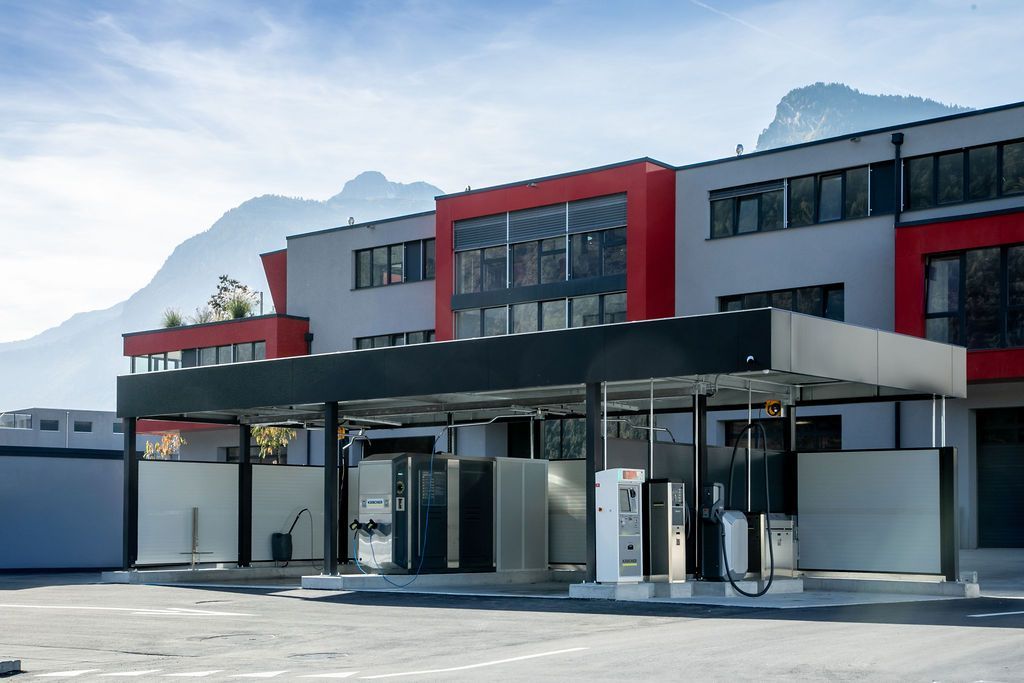 Station-service avec bâtiment gris et rouge, auvent au-dessus des pompes, montagnes en arrière-plan.