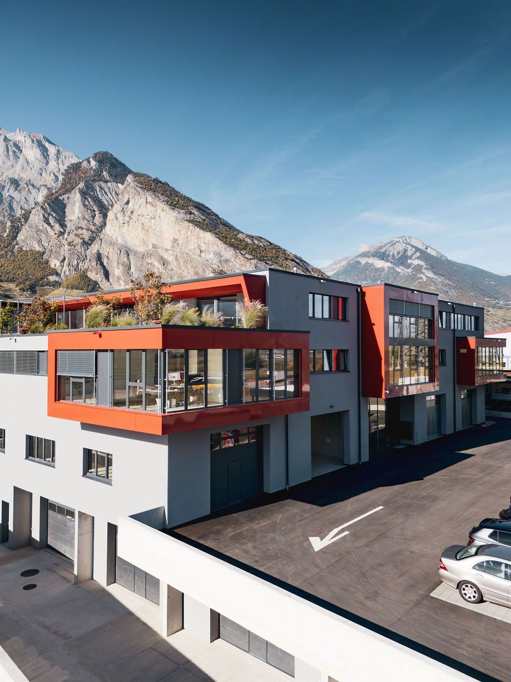 Bâtiment moderne avec des touches de rouge, vue sur les montagnes et parking.