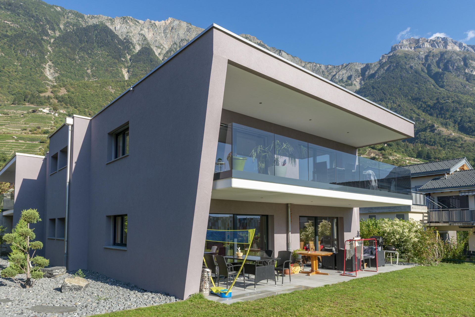 Maison grise moderne avec balcon vitré, terrasse et vue sur les montagnes.