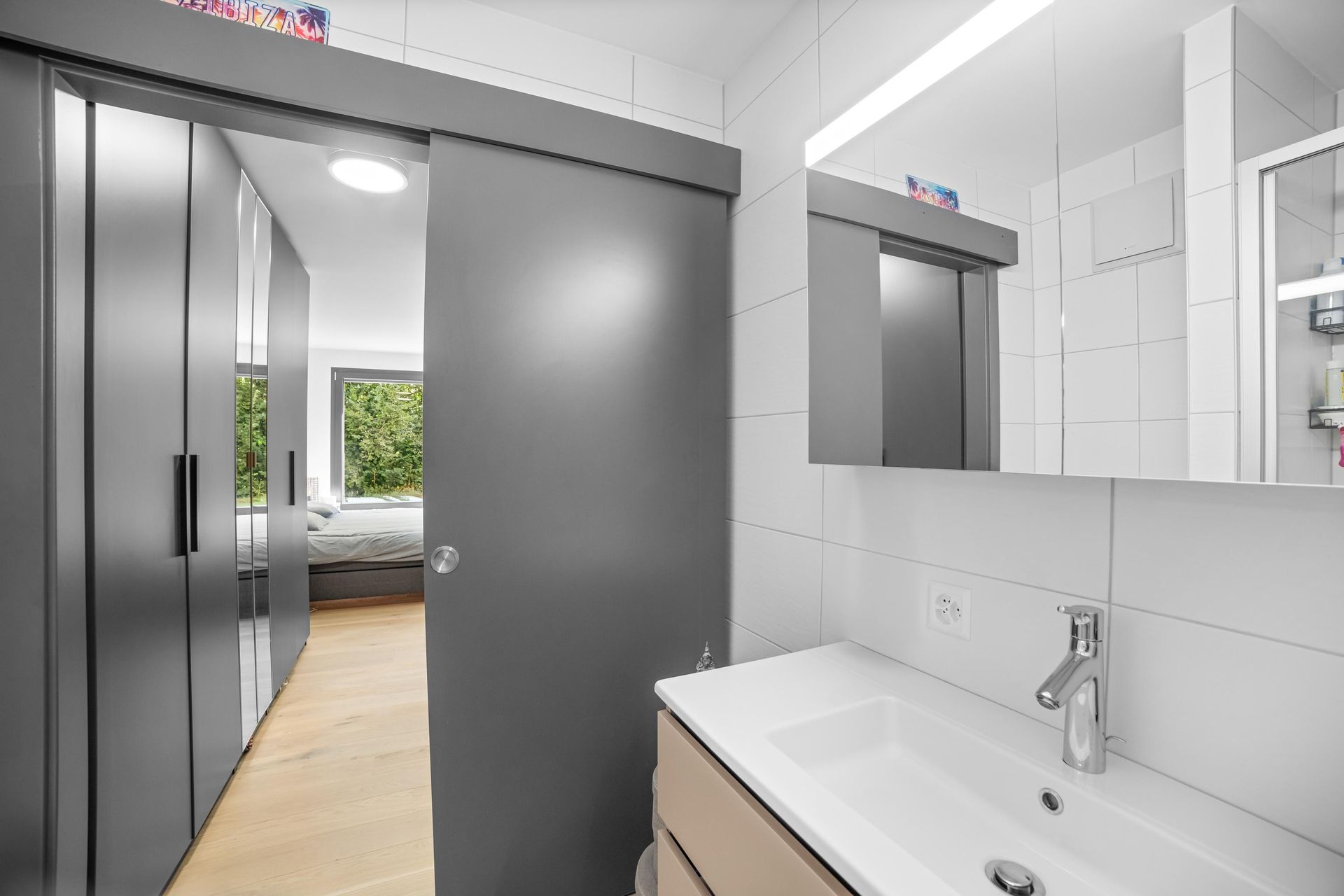 Une salle de bain avec lavabo, miroir et portes coulissantes donnant sur une chambre.