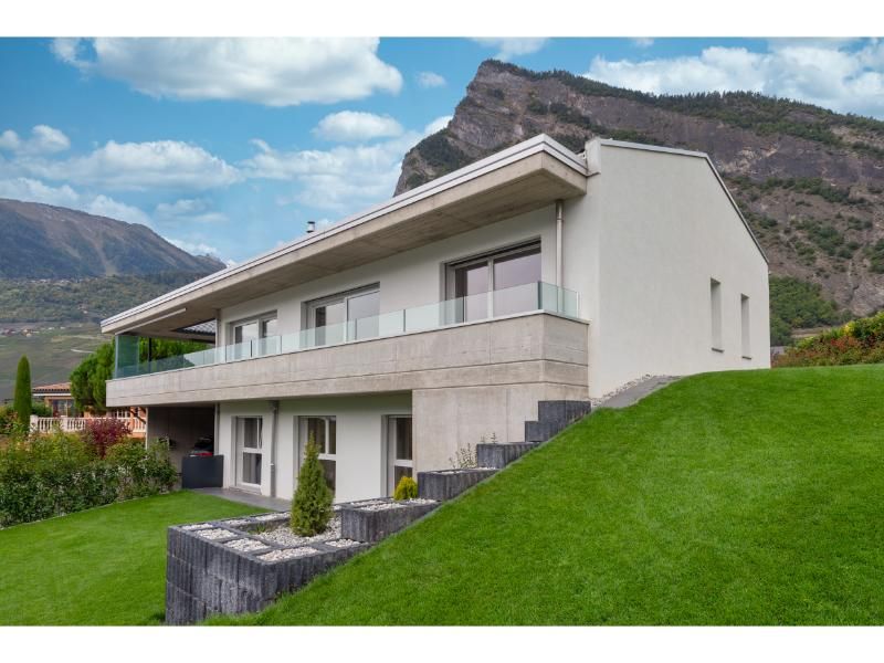 Une maison blanche est assise au sommet d'une colline herbeuse avec des montagnes en arrière-plan.