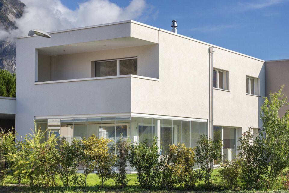 Une maison blanche avec beaucoup de fenêtres est entourée de buissons et d'arbres.