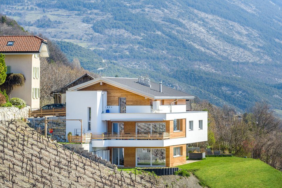 Maison blanche moderne avec des éléments en bois, située à flanc de colline, avec de l'herbe verte, un vignoble et des montagnes en arrière-plan.