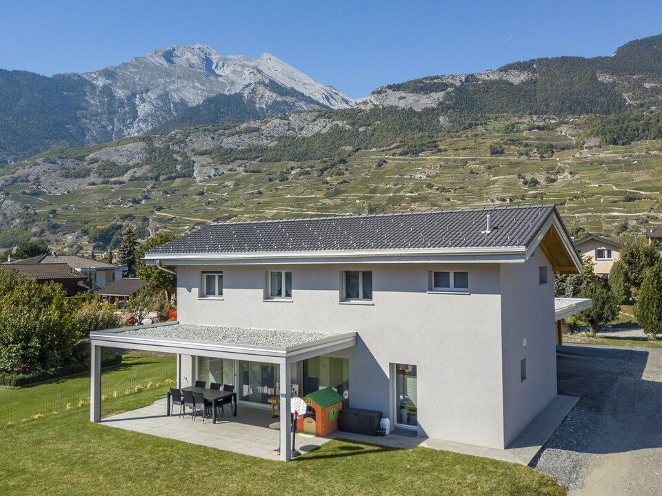 Maison à deux étages avec toit gris et terrasse, avec les montagnes en arrière-plan.