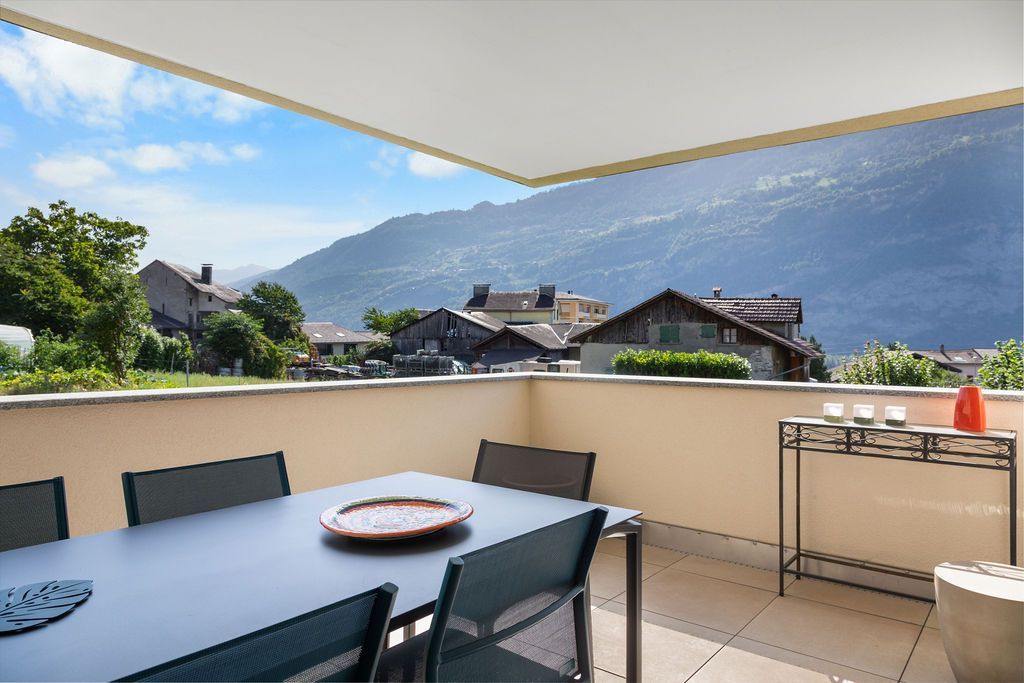 Terrasse avec table et chaises, donnant sur la ville et les montagnes. Ciel bleu.
