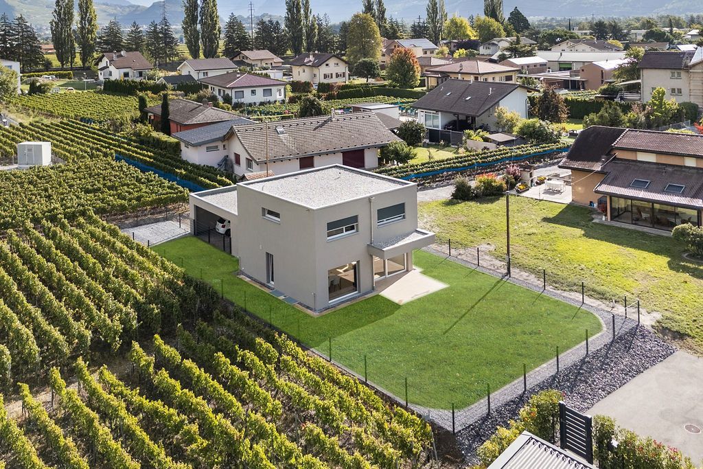 Maison moderne de deux étages avec toit plat, située au milieu d'un vignoble, entourée de pelouse et d'une petite clôture noire.