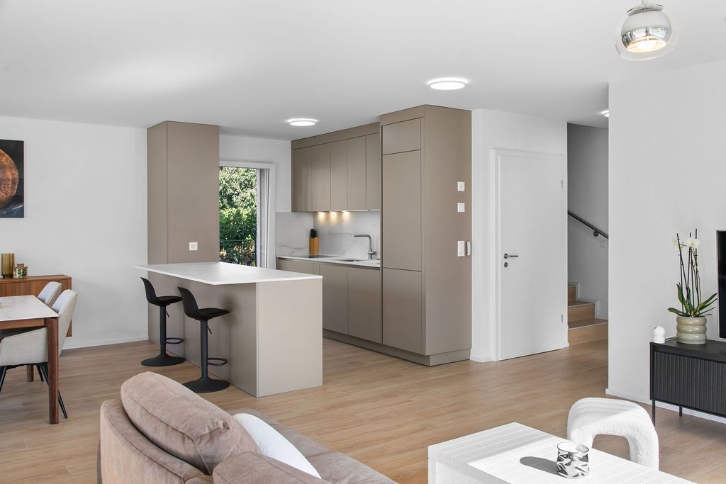 Espace de vie moderne et ouvert comprenant cuisine, coin repas et escalier. Placards beiges, parquet clair et murs blancs.