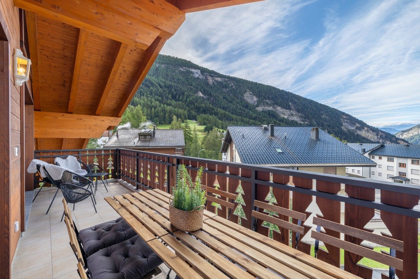 Balcon en bois donnant sur les montagnes et les toits ; on aperçoit une table, des chaises et une plante en pot.