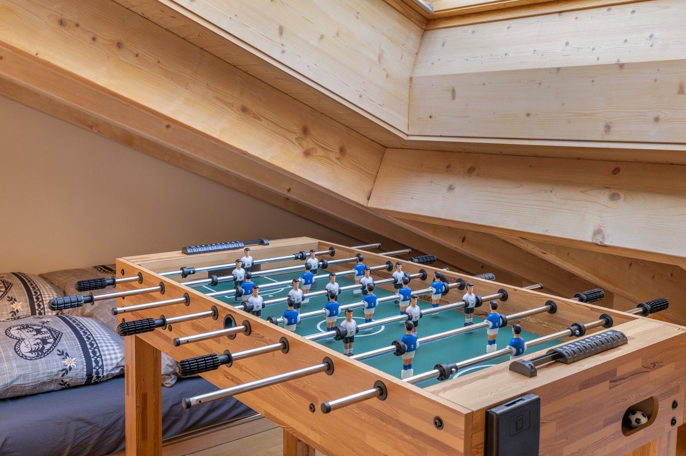Un baby-foot dans une chambre mansardée aux poutres apparentes, à côté d'un lit orné de coussins à motifs.
