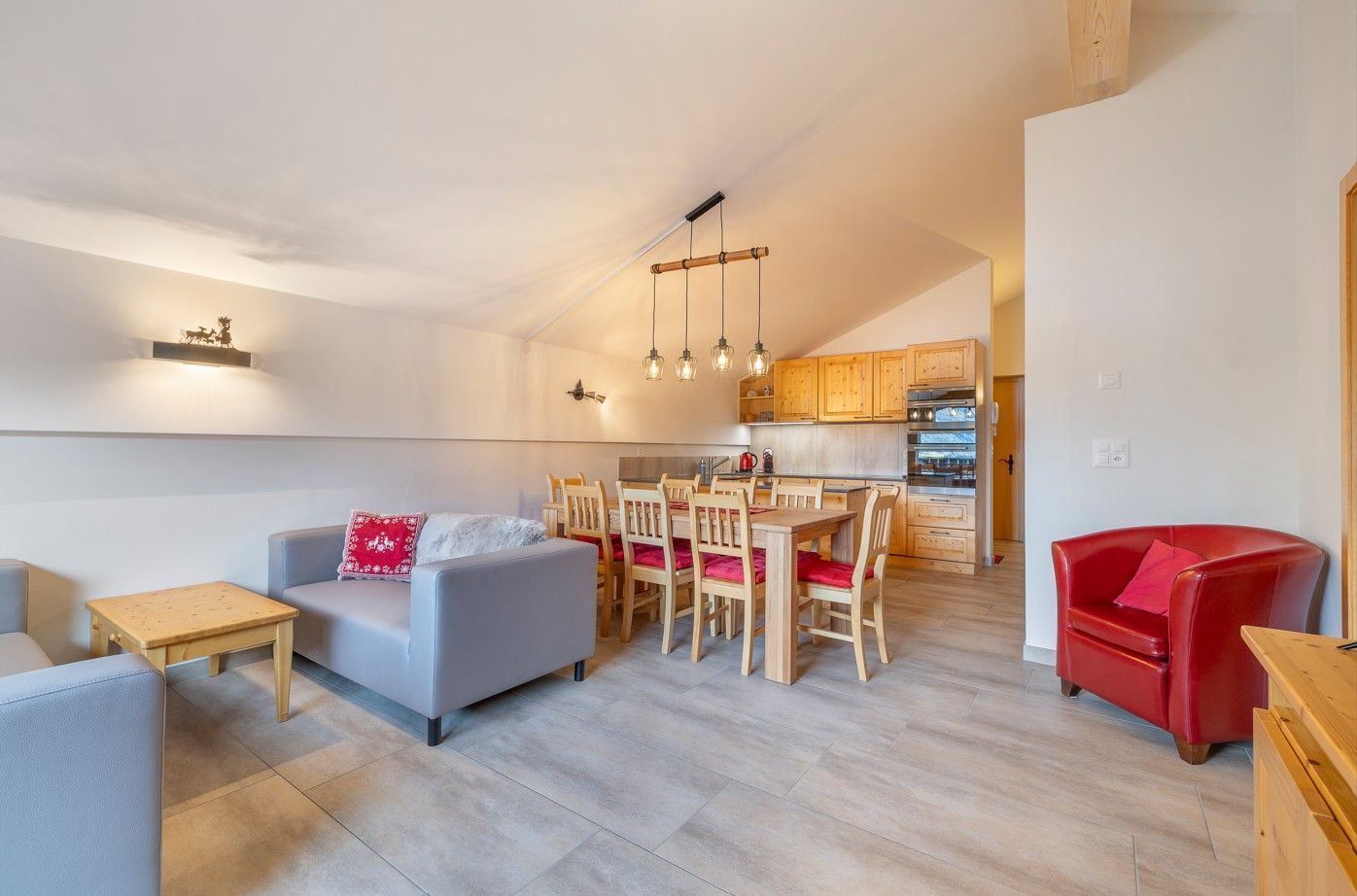 Vaste séjour comprenant un coin salon, une table à manger, une cuisine et un fauteuil rouge. Bois clair et murs blancs.